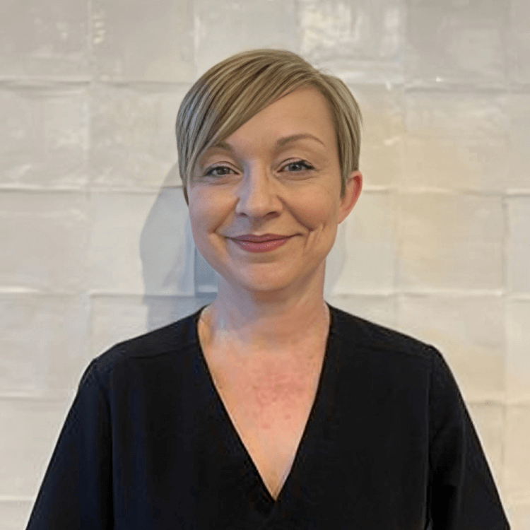 Smiling woman with short blonde hair in black top against white tiled background.