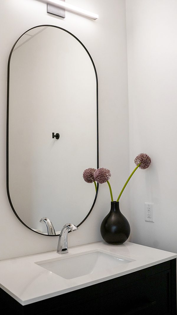 Minimalist black vase with purple flowers beside an arched mirror on a white wall.