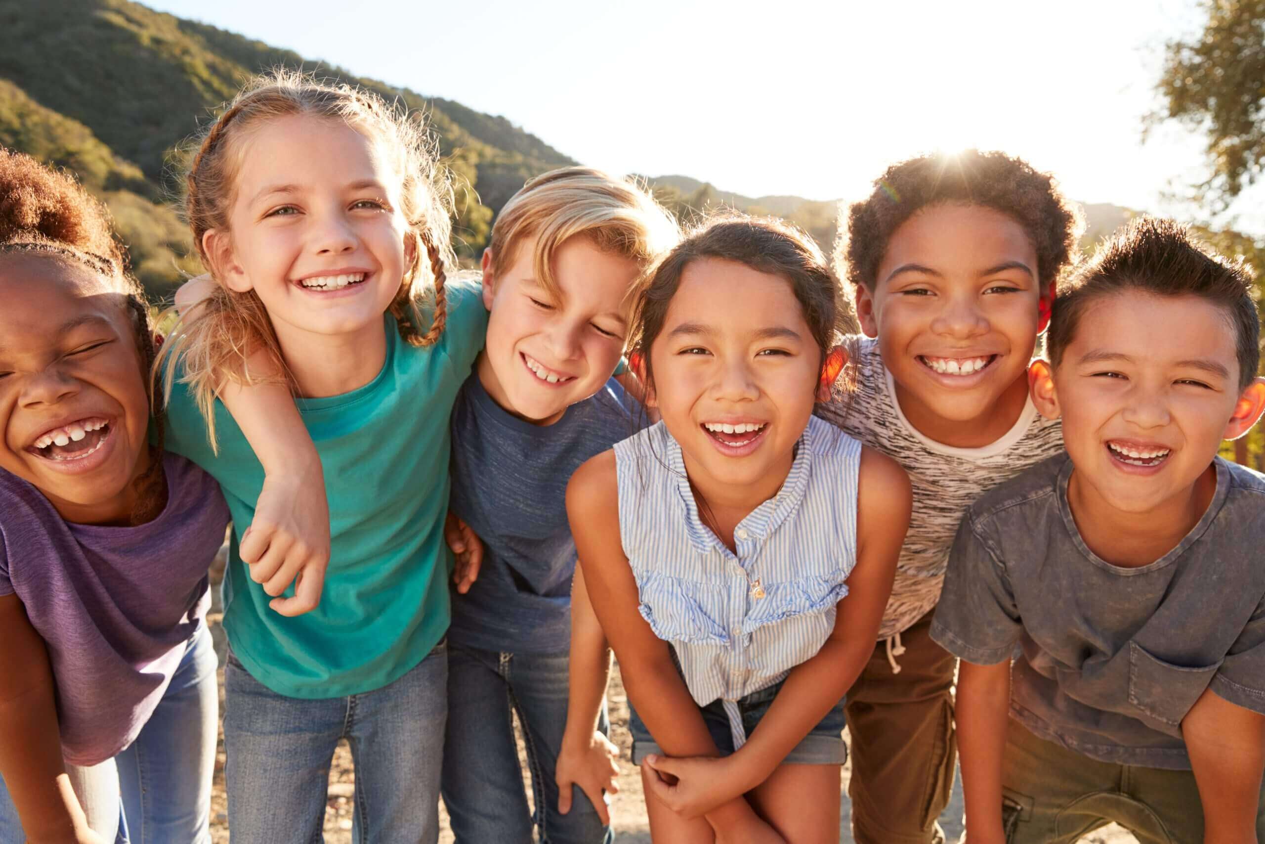 Four happy children smiling and hugging outdoors on a sunny day.